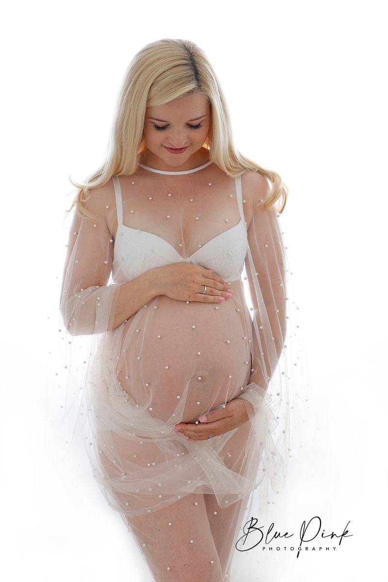 A pregnant woman in a flowing, transparent gown gently cradles her baby bump while looking down, with a radiant white background behind her, facing forward.